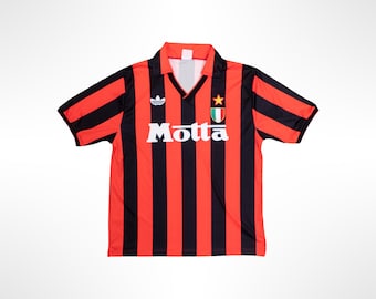 May include: A short-sleeved sports jersey featuring vertical red and black stripes, a V-neck collar, and a white Motta logo across the chest. The shirt includes a white Adidas trefoil logo on the right and a shield emblem with a star on the left.