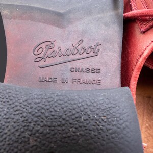 May include: The sole of a red leather shoe featuring the embossed text Paraboot, CHASSE, and MADE IN FRANCE. The heel has a textured black rubber surface, while the arch area is smooth and dark brown, showing signs of wear and use.