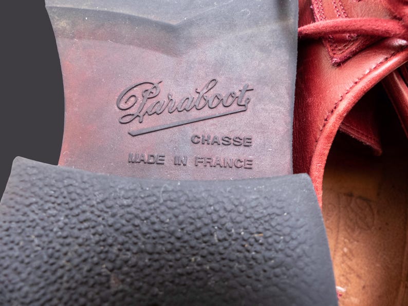 May include: The sole of a red leather shoe featuring the embossed text Paraboot, CHASSE, and MADE IN FRANCE. The heel has a textured black rubber surface, while the arch area is smooth and dark brown, showing signs of wear and use.