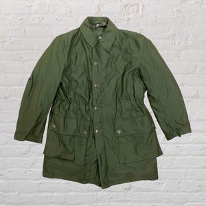 Puede incluir: Una chaqueta verde oliva con cierre de botones, cuello y cuatro bolsillos delanteros. La chaqueta tiene cintura con cordón y mangas largas. El fondo es una pared de ladrillos blancos.