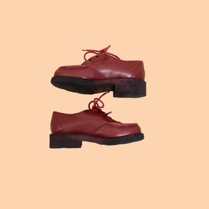 May include: A pair of dark red leather oxford shoes featuring a moccasin-style stitched toe, matching red laces, and a thick, dark brown rubber lug sole. The shoes have a low-cut silhouette with a rounded toe box and a sturdy, block-style heel.