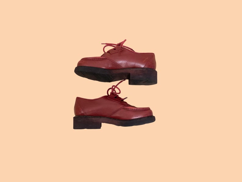 May include: A pair of dark red leather oxford shoes featuring a moccasin-style stitched toe, matching red laces, and a thick, dark brown rubber lug sole. The shoes have a low-cut silhouette with a rounded toe box and a sturdy, block-style heel.
