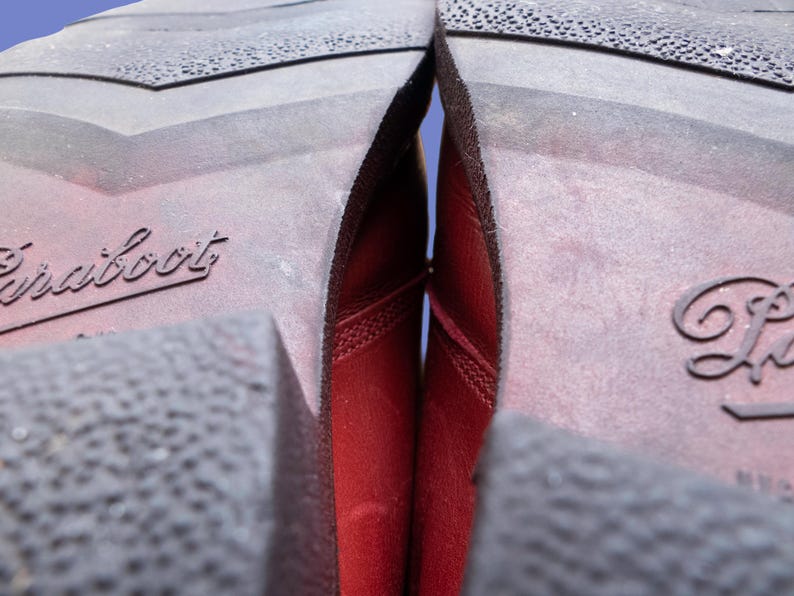 May include: The dark red leather uppers of a pair of boots are visible between the dark brown rubber soles. The soles feature a textured tread pattern and the word Paraboot is embossed on the heel area of each sole in a cursive script font.