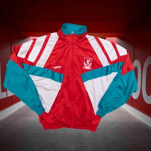 Könnte beinhalten: Rote, weiße und türkisfarbene Vintage-Adidas-Trainingsjacke. Die Jacke hat einen durchgehenden Reißverschluss, einen Stehkragen und das Wappen des Liverpool Football Club. Das Adidas-Logo ist auf der Brust sichtbar.