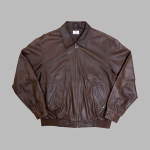May include: A brown leather jacket with a full zipper closure, two front pockets, and a collared neckline. The jacket has a ribbed hem and cuffs. The jacket is displayed on a gray background.