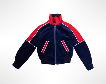 May include: A navy blue velour zip-up jacket featuring a high collar with two buttons, bright red panels along the shoulders and sleeves, and two front pockets with red and white striped trim. The jacket has a ribbed hem and sleeve cuffs.