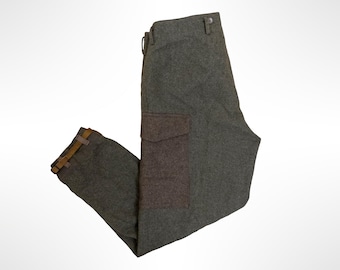 May include: Gray wool-blend trousers featuring a large dark brown cargo pocket on the side, a button closure at the waistband, and a brown adjustable strap with a metal buckle at the hem of the leg to secure the fit around the lower calf area.
