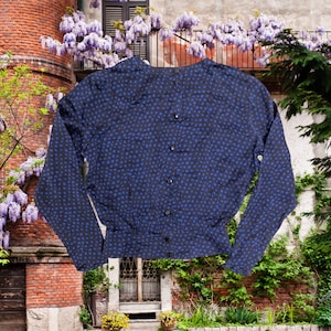 Könnte beinhalten: Marineblaue Bluse mit kleinen blauen Tupfen. Sie hat eine Knopfleiste vorne und eine geraffte Taille. Die langen Ärmel sind mit Knöpfen an den Manschetten versehen. Im Hintergrund ein Backsteingebäude und blühende Glyzinien.