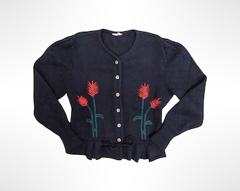 May include: A black knit cardigan featuring a round neckline, long sleeves with ribbed cuffs, and a button-down front. The garment is decorated with embroidered red and green floral motifs on the front panels and includes a ruffled hemline.
