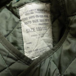 Puede incluir: Primer plano del interior de una chaqueta acolchada verde con una etiqueta cosida. La etiqueta contiene texto como "SMOCK MAN'S RAF GROUND TRADESMAN", "NATO SIZE 7090/0010" y "SIZE 190/112". El interior de la chaqueta tiene un patr&oacute;n de diamantes.