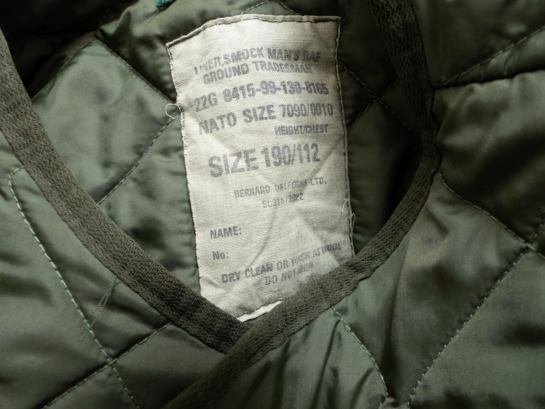 Puede incluir: Primer plano del interior de una chaqueta acolchada verde con una etiqueta cosida. La etiqueta contiene texto como "SMOCK MAN'S RAF GROUND TRADESMAN", "NATO SIZE 7090/0010" y "SIZE 190/112". El interior de la chaqueta tiene un patr&oacute;n de diamantes.
