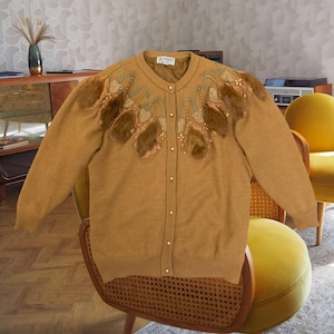 May include: Mustard-colored cardigan with decorative embellishments. The sweater features a round neckline, long sleeves, and a button-down front. The shoulders and upper chest are adorned with brown and gold decorative elements.