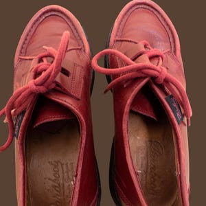 May include: A pair of red leather shoes with matching red laces and a moccasin-style toe construction. The interior insoles feature the embossed Paraboot brand name. The shoes have a dark sole and a small branded fabric tag on the side.