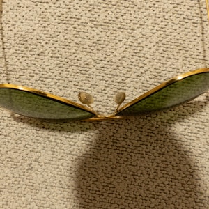 May include: Gold-rimmed sunglasses with green lenses. The sunglasses have a classic aviator shape, with a double bridge and thin gold arms. The lenses are a dark green color, and the frame is a shiny gold.