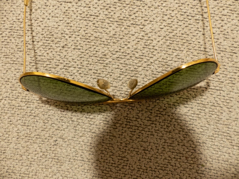May include: Gold-rimmed sunglasses with green lenses. The sunglasses have a classic aviator shape, with a double bridge and thin gold arms. The lenses are a dark green color, and the frame is a shiny gold.