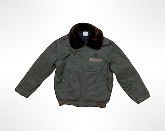 Puede incluir: Una chaqueta bomber de color verde oliva oscuro con un cuello de piel sintética marrón oscuro, cierre de cremallera frontal, mangas largas y un dobladillo acanalado marrón. La chaqueta incluye un detalle de parche rectangular en el pecho.