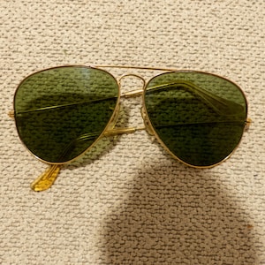 May include: Gold-framed aviator sunglasses with green lenses. The sunglasses have a double bridge and a classic teardrop shape. The temples are a light amber color. The lenses are a dark green color.