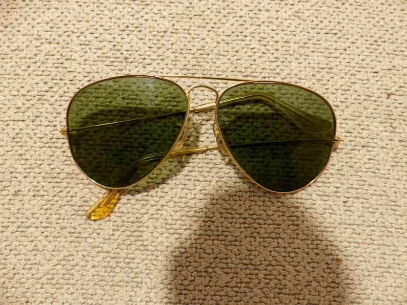 May include: Gold-framed aviator sunglasses with green lenses. The sunglasses have a double bridge and a classic teardrop shape. The temples are a light amber color. The lenses are a dark green color.