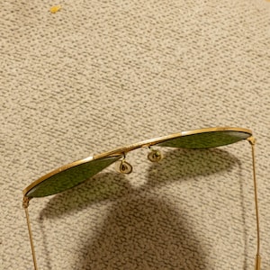 May include: Gold-rimmed sunglasses with green lenses. The aviator-style sunglasses have a double bridge and delicate gold arms. The lenses are a teardrop shape.