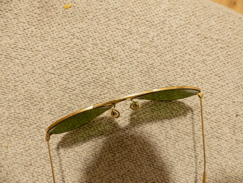 May include: Gold-rimmed sunglasses with green lenses. The aviator-style sunglasses have a double bridge and delicate gold arms. The lenses are a teardrop shape.