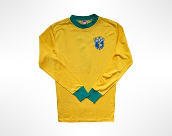 May include: A long-sleeve yellow shirt with a green crew neckline and green cuffs. The chest features a blue, yellow, and green shield-shaped emblem with the letters CBF and a small trophy graphic centered within the design.