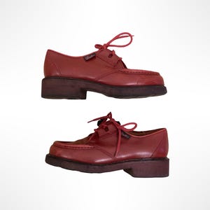 May include: A pair of red leather lace-up shoes featuring a moccasin-style stitched toe box, matching red laces, and a thick, dark brown rubber lug sole. The shoes have a low-cut silhouette with a small branded tag on the side of the eyelet panel.