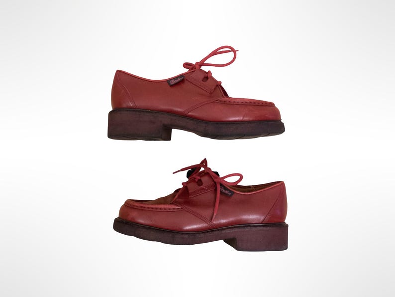 May include: A pair of red leather lace-up shoes featuring a moccasin-style stitched toe box, matching red laces, and a thick, dark brown rubber lug sole. The shoes have a low-cut silhouette with a small branded tag on the side of the eyelet panel.