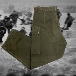 Può includere: Pantaloni cargo verde oliva con chiusura a bottone e tasca laterale. I pantaloni sono realizzati in un tessuto resistente e hanno un design classico e utilitario. I pantaloni sono mostrati su uno sfondo in bianco e nero.
