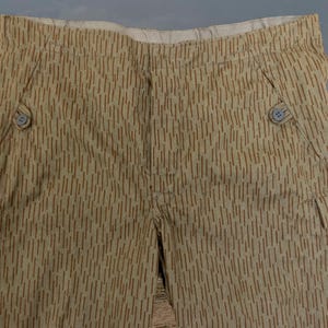 Puede incluir: Pantalones color beige con un patrón de rayas verticales marrones. Los pantalones tienen dos bolsillos delanteros, dos bolsillos laterales y dos botones en la parte delantera. La tela parece ligera.