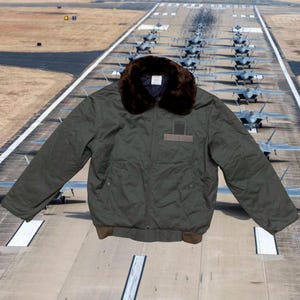 Könnte beinhalten: Olivgrüne Bomberjacke mit braunem Pelzkragen. Die Jacke hat einen Aufnäher auf der Brust und einen gerippten Saum. Im Hintergrund eine Landebahn mit Kampfflugzeugen.