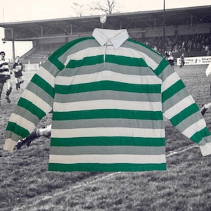May include: A rugby shirt with long sleeves, featuring green, white, and grey horizontal stripes. It has a white collar and cuffs. The background is a monochrome image of a rugby game in progress.