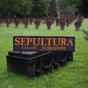 以下が含まれることがあります： オレンジ色の文字で「SEPULTURA CHAOS WORLD WIDE」と書かれた黒いバナーが、暗く装飾された石造りの構造物に置かれています。背景には、多数の石の十字架がある草地があります。