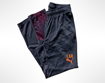 May include: Dark gray athletic pants featuring an elastic waistband and a tapered leg design. The lower left leg displays an orange Puma logo above the text LAR. The interior of the leg cuffs features a dark red and black geometric pattern.