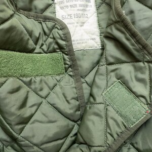 Puede incluir: Primer plano de un forro militar acolchado verde oliva. El forro presenta un patr&oacute;n de diamantes, dos parches de velcro verdes y una etiqueta con texto. Los bordes est&aacute;n ribeteados con un ribete verde oscuro.