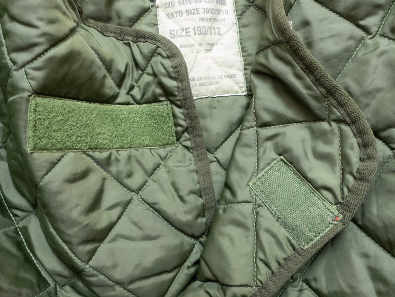 Puede incluir: Primer plano de un forro militar acolchado verde oliva. El forro presenta un patr&oacute;n de diamantes, dos parches de velcro verdes y una etiqueta con texto. Los bordes est&aacute;n ribeteados con un ribete verde oscuro.