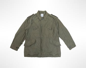 May include: An olive green field jacket featuring a pointed collar, long sleeves with buttoned cuffs, and two large flap pockets at the waist. The garment includes shoulder epaulets, a front button closure, and a visible white label at the inner neck.