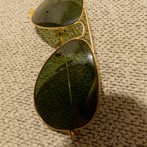May include: Gold-framed aviator sunglasses with green lenses. The sunglasses have a classic teardrop shape and a double bridge. The lenses reflect the surrounding environment. The frame is a shiny gold color.
