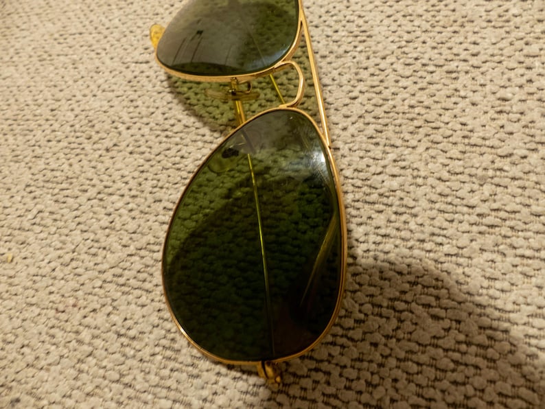 May include: Gold-framed aviator sunglasses with green lenses. The sunglasses have a classic teardrop shape and a double bridge. The lenses reflect the surrounding environment. The frame is a shiny gold color.