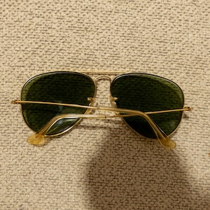 May include: Gold-framed aviator sunglasses with green lenses. The sunglasses have a classic teardrop shape and a thin gold metal frame. The temples and nose bridge are also gold. The lenses are a dark green color.