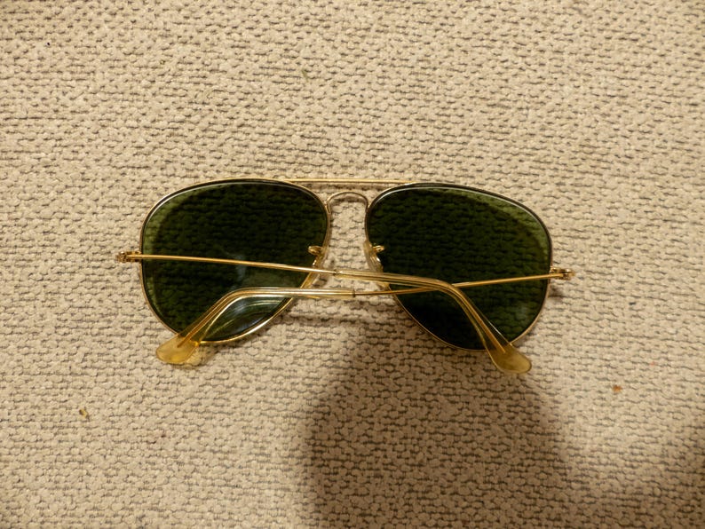 May include: Gold-framed aviator sunglasses with green lenses. The sunglasses have a classic teardrop shape and a thin gold metal frame. The temples and nose bridge are also gold. The lenses are a dark green color.