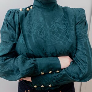May include: Teal green blouse with a high neckline and long sleeves. The fabric has a subtle, ornate pattern. Gold-colored buttons adorn the shoulders and cuffs. The blouse has a vintage aesthetic.