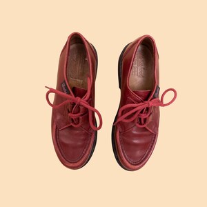 May include: A pair of dark red leather shoes with matching red laces and a moccasin-style stitched toe box. The shoes feature a low-cut silhouette, a small branded tag on the side, and a dark brown sole, presented from a top-down perspective.