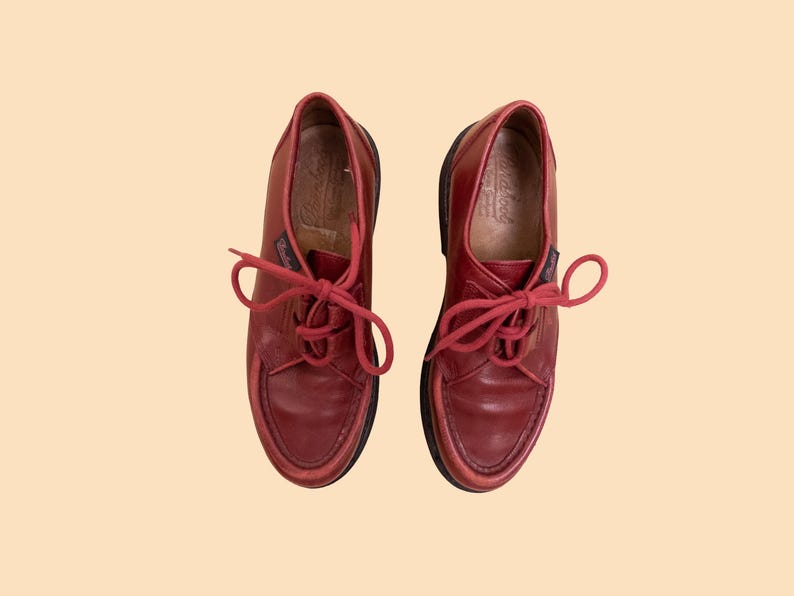 May include: A pair of dark red leather shoes with matching red laces and a moccasin-style stitched toe box. The shoes feature a low-cut silhouette, a small branded tag on the side, and a dark brown sole, presented from a top-down perspective.