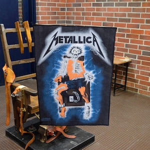 May include: Black rectangular Metallica band tapestry featuring a skeleton in an electric chair with lightning bolts. The band's logo is at the top. The tapestry is displayed in front of a wooden chair with leather straps.