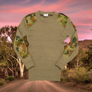 Peut inclure: Chemise à manches longues côtelée vert olive avec des patchs de camouflage sur les épaules et les coudes. La chemise a un col rond et une étiquette au niveau de l'encolure. Le motif de camouflage comprend des nuances de vert, de marron et de beige.