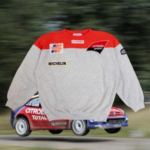 Peut inclure: Sweat-shirt gris avec épaules rouges et divers écussons, dont "CITROEN SPORT" et "MICHELIN". Le sweat-shirt est posé sur une voiture Citroën rouge et bleue.