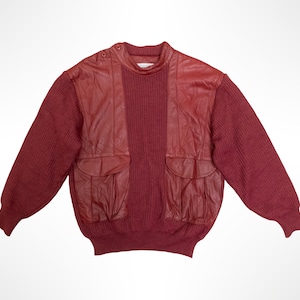 May include: A long-sleeve sweater in a deep red hue, featuring a central vertical knit panel flanked by smooth leather sections. The garment includes two front pockets with flaps, a ribbed crew neckline with button details, and ribbed cuffs.