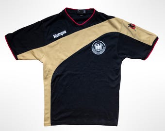 May include: A black and gold short-sleeve athletic jersey with a V-neck collar featuring red trim. The shirt displays the Kempa logo in white, a circular German Handball Federation emblem, and a small graphic of a handball player on the sleeve.