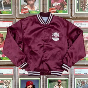 Peut inclure: Veste en satin bordeaux avec garniture rayée blanche et noire, avec le logo des Phillies. La veste est présentée sur une collection de cartes de baseball vintage. La veste mesure environ 76 cm de long.