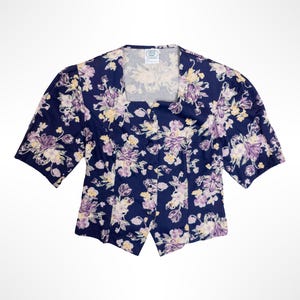 Op de afbeelding: Een marineblauwe blouse met korte mouwen, een vierkante halslijn en een bloemenpatroon in tinten lichtpaars, crème en geel. Het kledingstuk heeft een knoopsluiting aan de voorzijde, pofmouwen en een puntige zoom in het midden van de taille.